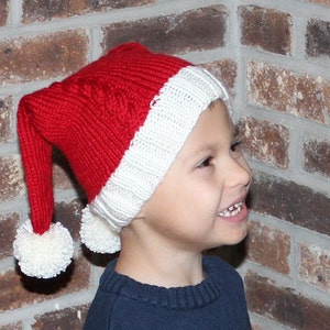 May include: A child wearing a red and white knitted Santa hat with white pom-poms. The hat has a long pointed tip.