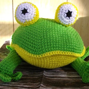 Op de afbeelding: Een groene gehaakte kikker kussen met gele accenten en grote zwarte ogen. De kikker zit op een houten oppervlak.