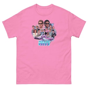 Op de afbeelding: Een felroze T-shirt met korte mouwen en een grafische print van de Miami Vice-cast, een sportwagen en een speedboot. Het ontwerp bevat een driehoekig achtergrondelement en de tekst MIAMI VICE in een gestileerd blauw en roze lettertype.