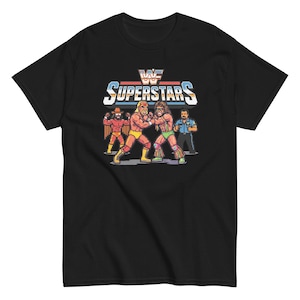 Può includere: Una t-shirt nera a maniche corte con una grafica colorata in pixel art raffigurante quattro lottatori. Sopra le figure, il testo recita WWF SUPERSTARS in un carattere audace e stilizzato con strisce orizzontali rosse, bianche e blu dietro.