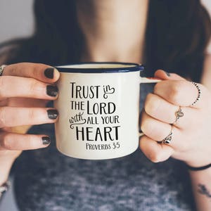 Może przedstawiać: Kremowy emaliowany kubek z niebieską obw&oacute;dką i uchwytem, z napisem "Trust in the Lord with all your heart" i wersetem biblijnym Przypowieści 3:5 w czarnym kolorze. Kubek jest trzymany przez osobę.