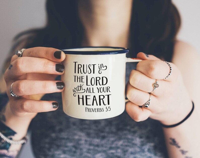 Może przedstawiać: Kremowy emaliowany kubek z niebieską obw&oacute;dką i uchwytem, z napisem "Trust in the Lord with all your heart" i wersetem biblijnym Przypowieści 3:5 w czarnym kolorze. Kubek jest trzymany przez osobę.