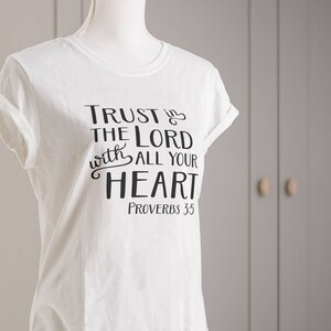 Może przedstawiać: Biała koszulka z podwiniętymi rękawami, z czarnym napisem "TRUST IN THE LORD with ALL YOUR HEART" i "PROVERBS 3:5". Koszulka jest prezentowana na manekinie na neutralnym tle.