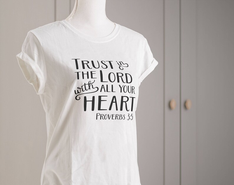 Może przedstawiać: Biała koszulka z podwiniętymi rękawami, z czarnym napisem "TRUST IN THE LORD with ALL YOUR HEART" i "PROVERBS 3:5". Koszulka jest prezentowana na manekinie na neutralnym tle.