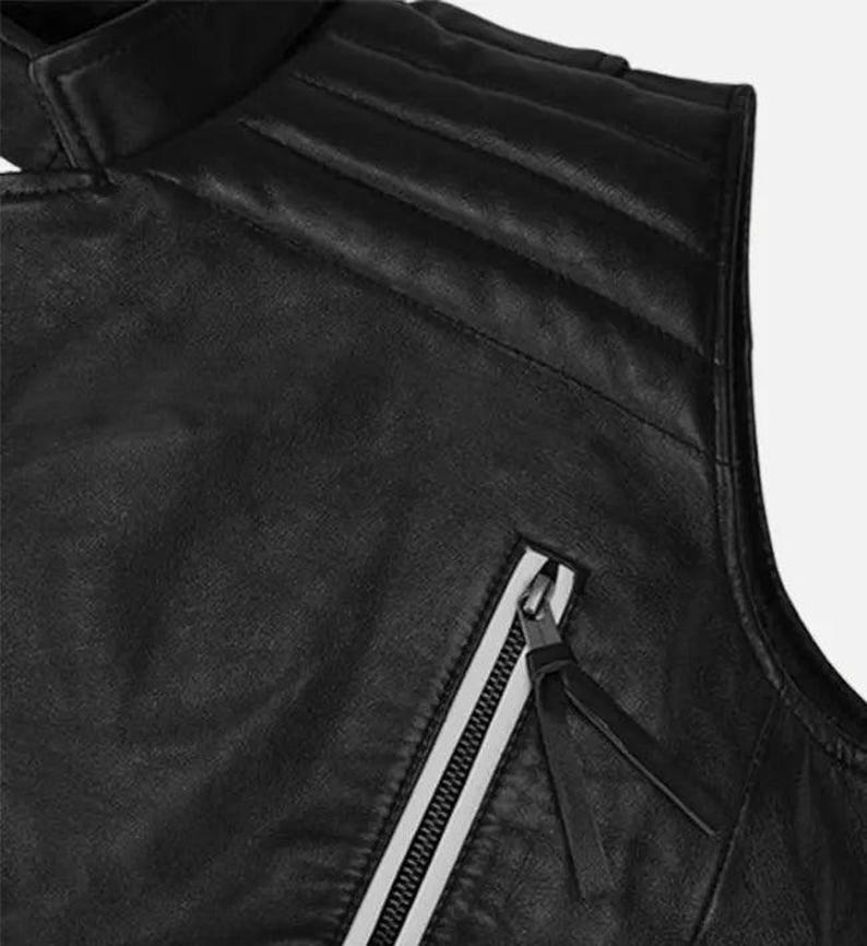 May include: Close-up of a black leather vest with a high collar and quilted shoulder detailing. The vest features a diagonal zipper with a white stripe and a black leather pull.