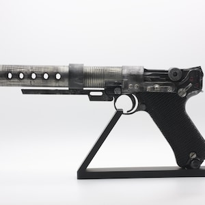 Op de afbeelding: Een blasterpistool-rekwisiet met een lange, metallic zilveren loop met ronde uitsparingen en een felrode punt. Het handvat is zwart met een getextureerde grip en de behuizing heeft een verweerde, donkere metallic afwerking op een zwarte standaard.