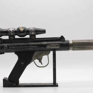 Op de afbeelding: Een zwart en metaalgrijs speelgoedblastermodel met een gemonteerde richtkijker, een getextureerde handgreep, een trekker met beugel en een cilindrische loop die uitloopt in een rode punt, geplaatst op een zwarte standaard.