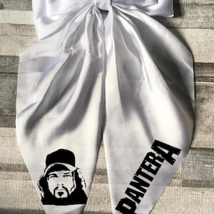 May include: White satin hair bow with a large bowknot. The bow tails feature a black and white image of a man wearing a baseball cap and the word "PANTERA" in bold black letters.