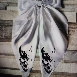 Op de afbeelding: Een witte satijnen haarstrik met een grote strikknoop en lange, vloeiende staarten. Elke staart heeft een zwarte afbeelding van een gestileerde schedel. De strik wordt waarschijnlijk gebruikt als haaraccessoire.