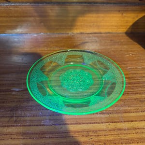 May include: A small, round, translucent neon green glass saucer featuring a detailed embossed floral pattern in the center and a decorative border along the rim. The glass exhibits a vibrant, glowing appearance characteristic of uranium glass.