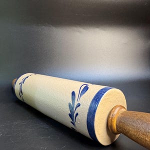 May include: A cream-colored rolling pin with blue floral designs and a dark wooden handle. The rolling pin has a blue band around the edge. The rolling pin is approximately 12 inches long.