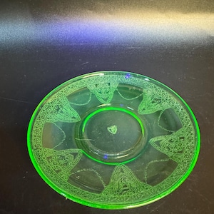 May include: A round, translucent neon green glass saucer featuring a decorative border with repeating arched patterns and intricate floral motifs. The center of the saucer displays a small, embossed shield-shaped emblem in the middle of the basin.