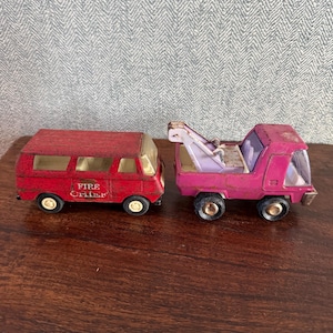 May include: A red toy van with the text FIRE CHIEF printed on the side and a pink toy tow truck with a white crane arm. Both vehicles feature weathered paint, clear plastic windows, and black wheels with white centers, displayed side by side.