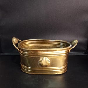 May include: An oval brass planter featuring horizontal ribbed detailing along the sides and a centered embossed seashell motif. The vessel includes two small, curved handles on opposite ends, providing a polished metallic finish throughout.