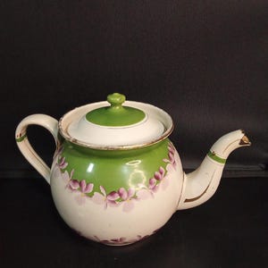 May include: A white enamel teapot featuring a green band around the upper body and lid. The teapot is decorated with a hand-painted floral pattern of small purple flowers encircling the middle. It has a curved handle and a spout with gold trim.