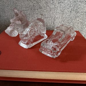Könnte beinhalten: Zwei klare Glasfiguren: ein Scottish Terrier Hund und ein Oldtimer. Der Hund steht, und das Auto hat detaillierte Merkmale. Beide stehen auf einem roten Buch.