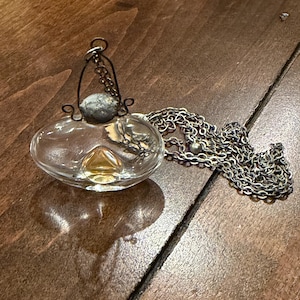 May include: A clear glass perfume bottle pendant on a silver-toned metal chain. The bottle has a rounded, flattened shape with a small amount of amber-colored liquid inside. A dark, textured metal cap is secured with a wire loop attached to the chain.