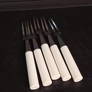 May include: A set of five two-pronged cocktail forks featuring metallic silver-toned heads and smooth, cylindrical white handles. The forks are arranged in a parallel row, showcasing the contrast between the polished metal tines and the solid white grips.