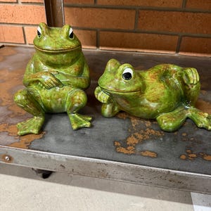 May include: Two green ceramic frog ornaments. One frog is seated with its arms crossed, the other is reclining. Both have white eyes and brown detailing. These frog sculptures are suitable for display in a garden or home. Each frog is a unique decorative item.