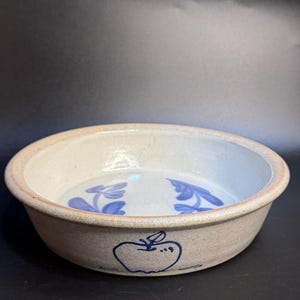 Puede incluir: Un cuenco redondo de gres con exterior beige claro e interior blanco. El cuenco presenta diseños de manzanas y hojas azules. El borde es de color beige más oscuro. El cuenco es adecuado para servir alimentos.