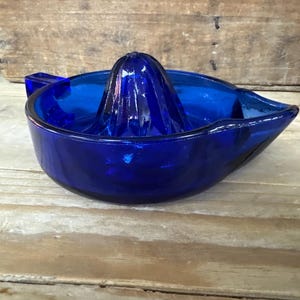 May include: A cobalt blue glass citrus juicer. The juicer features a ridged reamer in the center, surrounded by a bowl with a pouring spout. The juicer is made of thick, durable glass and is designed for easy use and cleaning.
