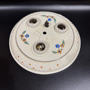 May include: Off-white ceramic ceiling light fixture with four bulb sockets. The fixture is decorated with blue and orange floral patterns and a border of leaves and orange dots. A central metal piece is visible.