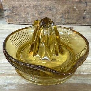Könnte beinhalten: Bernsteinfarbene Glas-Zitruspresse mit Reibahle und Schüssel. Die Schüssel hat ein geripptes Design auf der Innenseite. Die Zitruspresse besteht aus Glas und ist zum Auspressen von Zitrusfrüchten bestimmt. Die Zitruspresse steht auf einer Holzoberfläche.