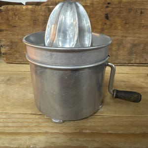 May include: A vintage, silver-toned citrus juicer with a ribbed reamer and a cylindrical base. It features a handle with a dark wooden grip and three small feet. The juicer is designed for manual use and is likely made of metal.