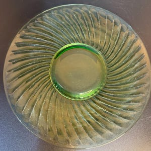 May include: A round, translucent green glass saucer featuring a central circular indentation for a cup. The outer rim of the saucer is decorated with a series of raised, radiating swirl patterns that extend from the center toward the edge.