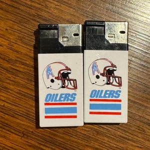 May include: Two white lighters with black tops. Each lighter features a football helmet graphic with the word "OILERS" in blue text. Below the text are blue and red stripes. The lighters are on a wooden surface.