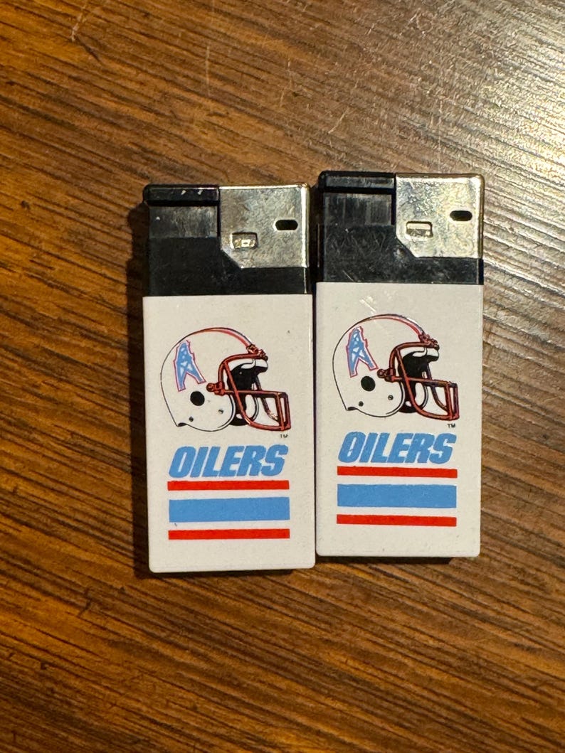 May include: Two white lighters with black tops. Each lighter features a football helmet graphic with the word "OILERS" in blue text. Below the text are blue and red stripes. The lighters are on a wooden surface.