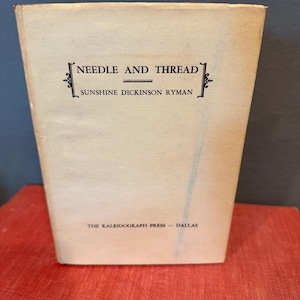 May include: A vintage book titled "Needle and Thread" by Sunshine Dickinson Ryman. The cover is a light beige colour with black text and decorative elements. The book is set on a red surface.
