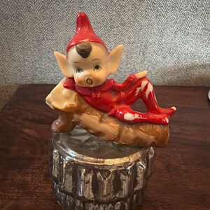 May include: A vintage ceramic elf figurine in a red outfit and hat, reclining on a log. The elf has a white face, large ears, and is resting on a silver-coloured container. The figurine is a decorative item.