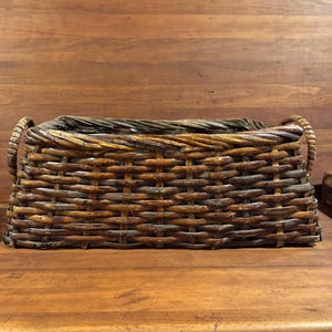 May include: A rectangular woven basket with curved handles. The basket is made of brown and tan wicker, with a textured surface. The basket is sitting on a wooden surface.