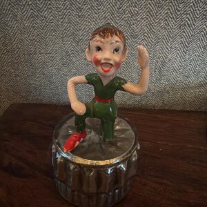 Könnte beinhalten: Vintage Peter Pan-Figur in grünem Outfit mit roter Feder an einem Fuß, stehend auf einem dekorativen Glasbehälter. Die Figur hat einen lächelnden Ausdruck und steht vor einem gemusterten Hintergrund.