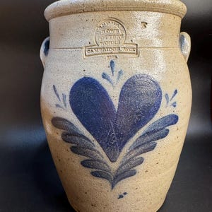 May include: A stoneware crock with a blue heart design and the text "ANDREW ROWE POTTERY WORKS CAMBRIDGE, WIS." The beige-coloured crock has two small handles and a wide opening. The heart design is surrounded by blue leaf-like accents.