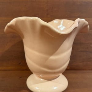 Pode incluir: Um vaso de cerâmica cor de pêssego com uma borda com babados. O vaso tem uma base de pedestal e uma forma de tulipa. A superfície tem uma textura sutil e rodopiante. O vaso é provavelmente vintage e adequado para exibir flores.