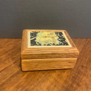 Op de afbeelding: Een kleine houten doos met een decoratief deksel. Het deksel heeft een kleurrijke illustratie met gele, blauwe en groene accenten. De doos is gemaakt van lichtgekleurd hout en heeft een rechthoekige vorm. De doos is waarschijnlijk voor het opbergen van kleine items.