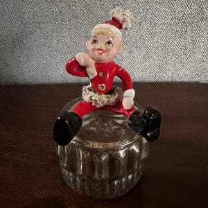 May include: A vintage Christmas figurine of a seated elf in a red suit with a white collar and hat, perched atop a clear, decorative glass container. The elf has a smiling face and black boots. The container has a ribbed design.