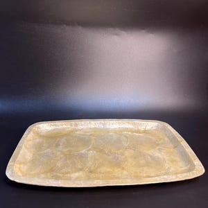 May include: A rectangular serving tray with rounded corners. The tray has a pearlescent, iridescent finish with a subtle, textured surface. The tray is a light, neutral colour, and is set against a dark background.