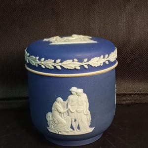 May include: A cylindrical blue stoneware jar with a matching lid, featuring white relief ornamentation. The body displays a classical scene of three figures, while the lid edge is decorated with a white leaf garland and a central relief motif.