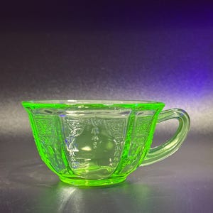 Può includere: Una tazza da tè in vetro verde brillante con manico. La tazza presenta un motivo decorativo attorno al bordo e ai lati. La tazza da tè è realizzata in un materiale di vetro traslucido.