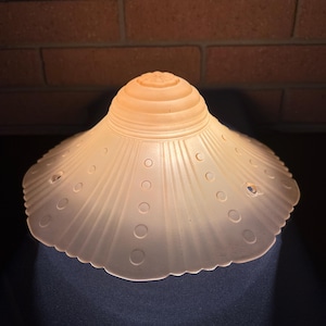 May include: A vintage, frosted glass lampshade with a scalloped edge. The shade has a conical top and a flared, circular bottom. It features a series of embossed circles and vertical lines radiating from the center. The color is a soft, warm white.