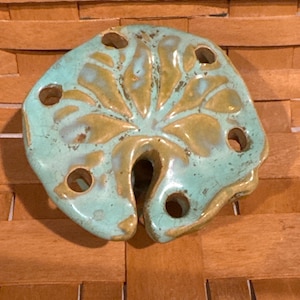 May include: A ceramic teal and gold leaf-shaped object with multiple holes. The object has a distressed finish and is likely a decorative item or a functional piece for home decor. The background is a woven basket.