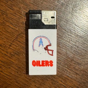 May include: A black and white lighter with a white front panel. The panel features a logo with a football helmet and an oil derrick, with the word "OILERS" in red block letters.