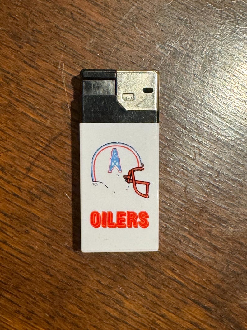 May include: A black and white lighter with a white front panel. The panel features a logo with a football helmet and an oil derrick, with the word "OILERS" in red block letters.