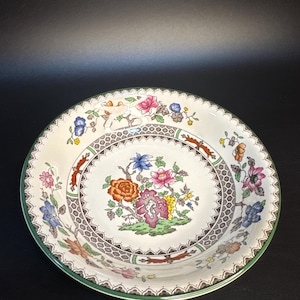 May include: A white ceramic bowl with a green rim and a floral design. The center of the bowl features a bouquet of colorful flowers, including pink, orange, and blue blooms, surrounded by a brown and white patterned border.