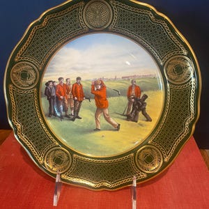 May include: A decorative plate with a green and gold border featuring a golf scene. The plate's border has a repeating chain design and geometric patterns. The central image depicts golfers on a green field under a blue sky.