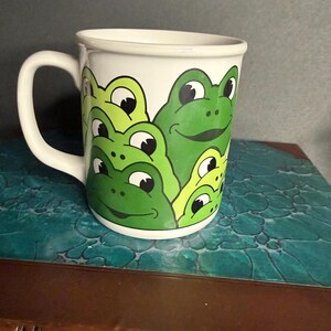 May include: White ceramic mug featuring a design of several green cartoon frogs with black eyes. The frogs are in various sizes and overlapping each other. The mug has a white handle and a white rim.