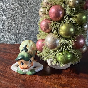 Puede incluir: Una figura de cerámica vintage de un elfo sonriente con sombrero y atuendo verdes, apoyado en los codos. Un pequeño árbol de Navidad con agujas verdes y adornos en tonos rosa, dorado y verde en el fondo. Los objetos están sobre una superficie de madera.